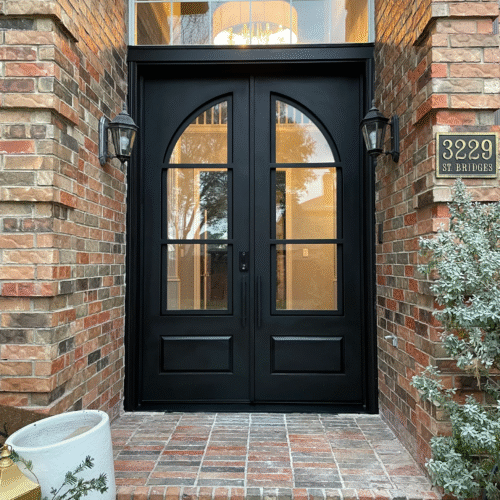 Custom black double iron door with arched glass panels installed on brick home